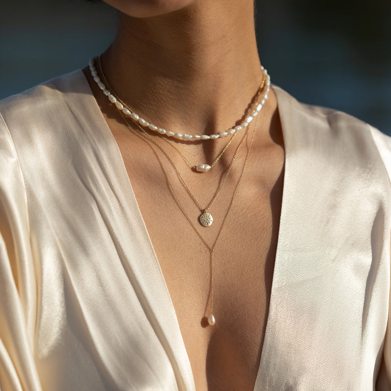 A woman wearing a silky, cream-colored blouse is adorned with three layered necklaces. The necklaces include a choker with white beads, a pendant necklace with a round, textured charm, and the sofia perla necklace - 14k yellow gold necklace with pearl by bluboho.