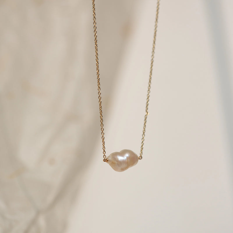 The sofia perla necklace by bluboho, a 14k yellow gold chain necklace, showcases a single, uniquely shaped pearl pendant. The soft and neutral background highlights the jewelry piece's simplicity and elegance.