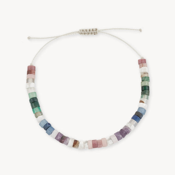 The bluboho rainbow bead bracelet features an array of cylindrical gemstone beads in pastel shades of pink, green, blue, purple, and white. These colorful beads are strung on a beige cord with an adjustable sliding knot closure, and the bracelet is showcased in a circular shape against a plain white background.