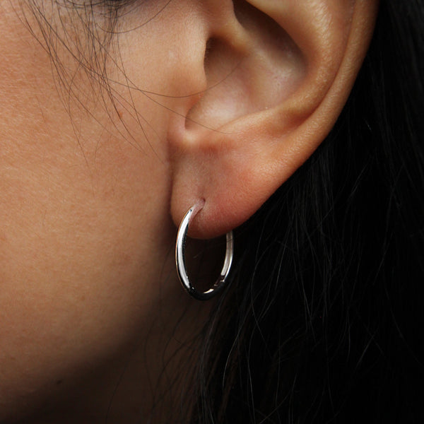 Close-up of a person's ear wearing the bluboho dagger large hoop silver earring. Dark hair is partially visible around the ear, and the background is out of focus.