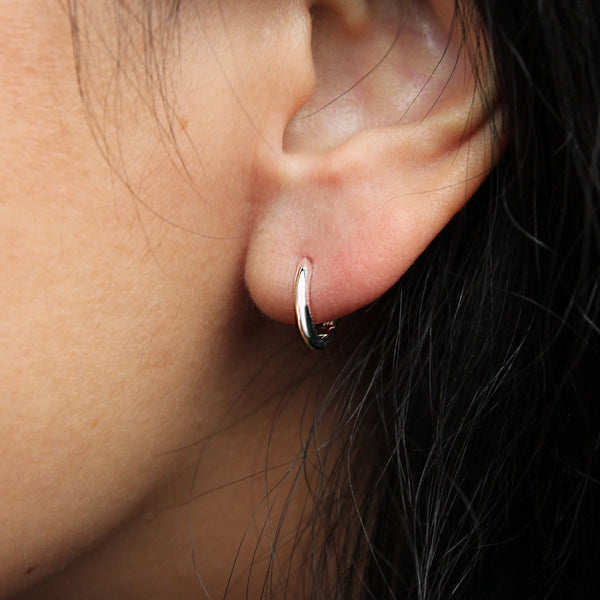 Close-up of a person's ear wearing the bluboho dagger small hoop silver earring. The simple and elegant earring features a smooth, shiny finish. The person's hair is partially visible, cascading against their ear and neck, contrasting with their light skin tone.