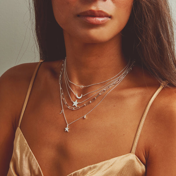 A person wearing a satin spaghetti strap top is shown from the shoulders up. They are adorned with multiple delicate silver necklaces, including the Supernova White Sapphire Necklace in sterling silver by bluboho, featuring charms like stars, moons, and beads. The background is a plain, neutral color.