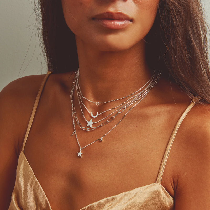 A person wearing a beige spaghetti strap top showcases multiple layered silver necklaces. The necklaces feature various charms including a star, a crescent moon, a tiny star, and geometric shapes, creating a delicate and stylish look. One of the standout pieces is the "Bright Star White Topaz Baguette Necklace" in sterling silver by bluboho. The background is plain and neutral.