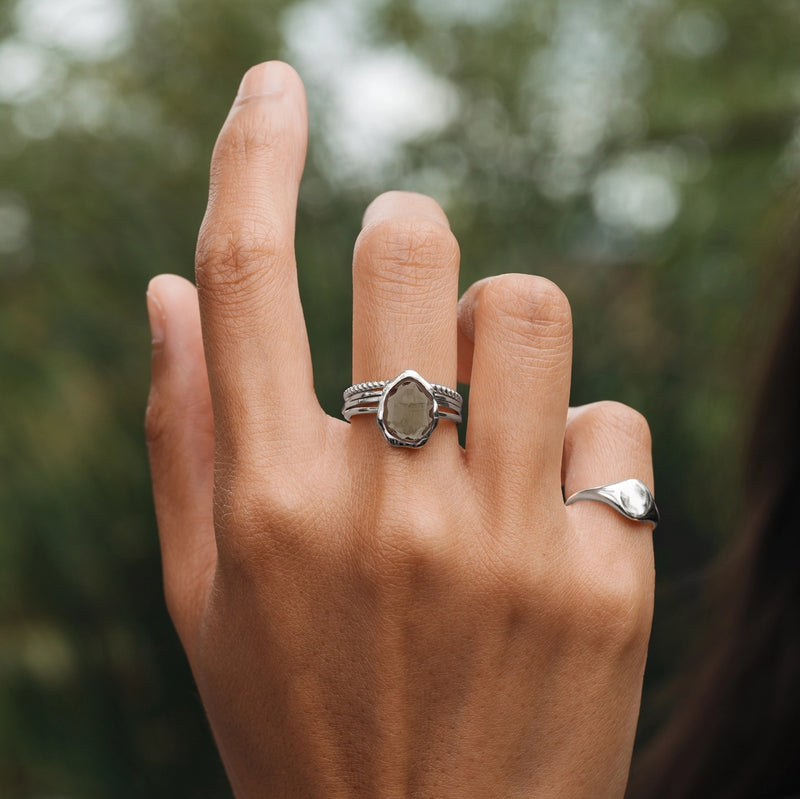 Close-up of a hand raised against a blurred green background, showcasing two rings on the middle finger. The main ring, named the "smoky quartz mood ring silver" by bluboho, features an irregular smoky quartz stone encased in sterling silver with a detailed band. The smaller ring on the pinky has a minimalist, smooth silver design.