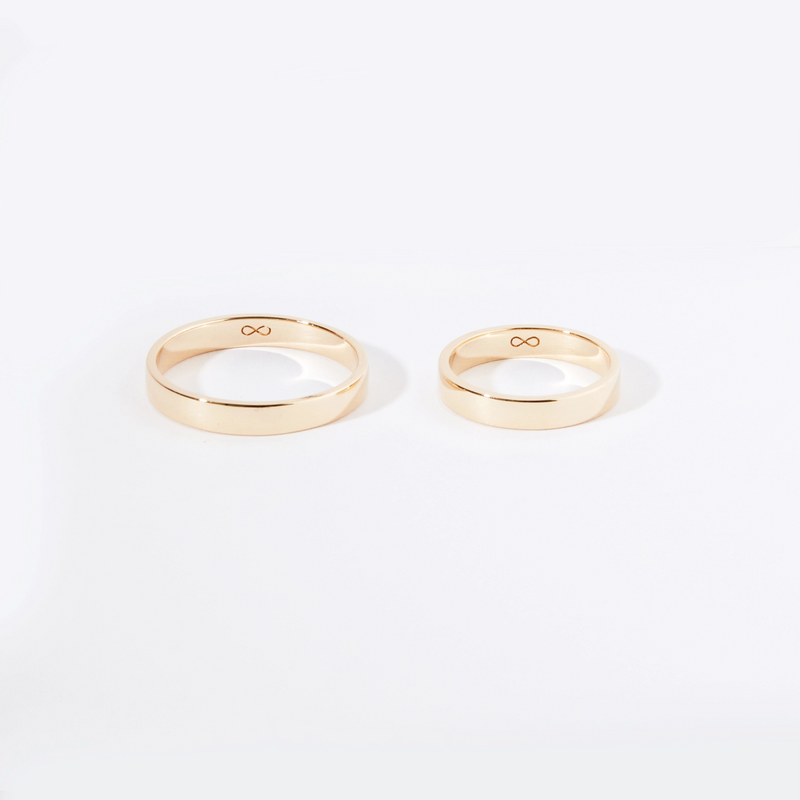 Two gold wedding bands on a white background. The rings are simple and polished, with an engraved infinity symbol on the inner side of each band. One ring is slightly larger than the other, suggesting a pair meant for couples. These are the "infinity love band polished - 14k yellow gold" by "beloved by bluboho.