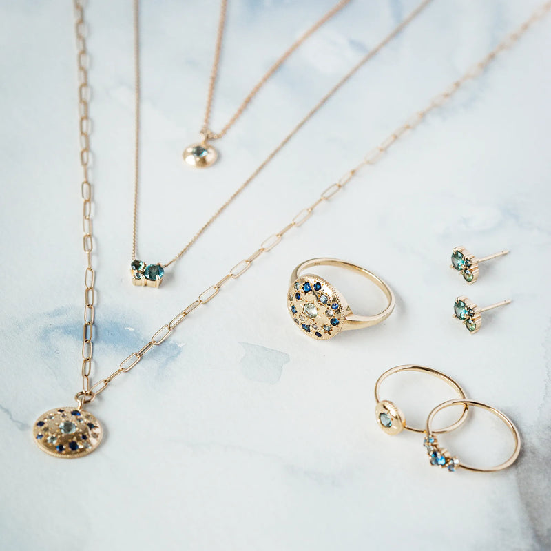 A collection of gold jewelry from bluboho is displayed on a light blue and white marble background. The set includes the "something blu tie dye moonchild ombre sapphire necklace" in yellow gold, along with rings and matching stud earrings featuring blue and green stones.