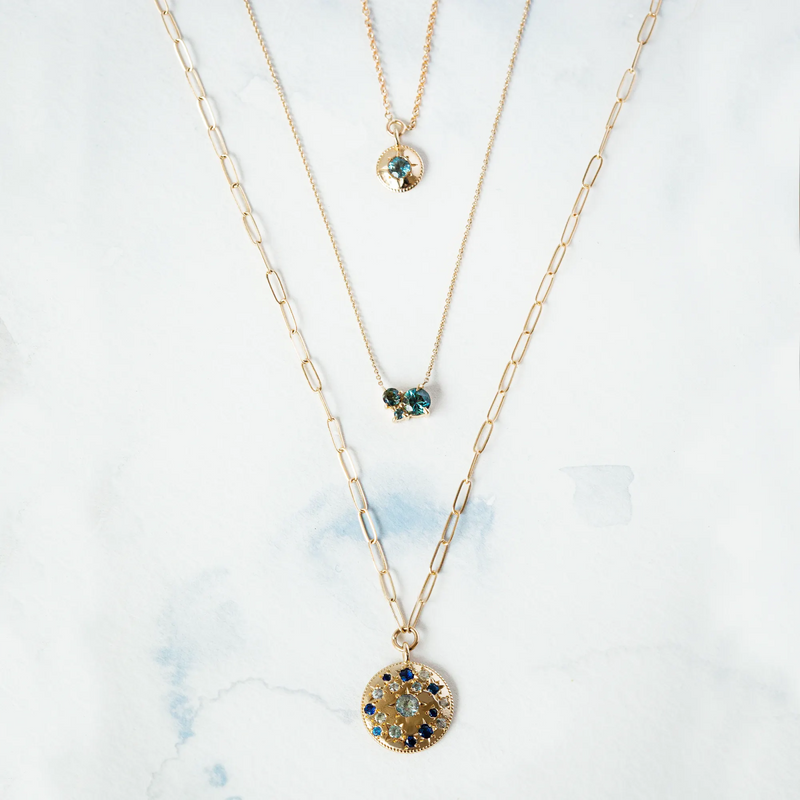 Three gold necklaces are arranged vertically on a light surface. The shortest necklace features a small pendant with blue gemstones, the middle necklace showcases a butterfly-shaped pendant, and the longest one is the "something blu tie dye moonchild ombre sapphire necklace" by bluboho, featuring round pendants adorned with sapphires.