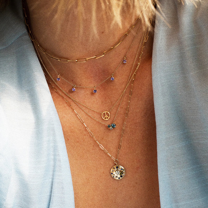 A person adorned with the bluboho something blu tie dye moonchild ombre sapphire necklace in yellow gold, featuring an intricate arrangement of charms: a peace symbol, a small round pendant set with multicolored stones, small blue and purple gems, and a chain-link design. They are wearing a light blue blouse that is slightly open.