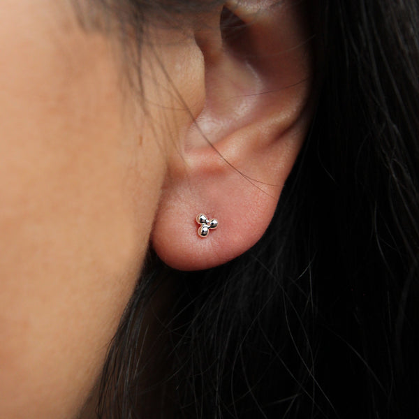 Close-up of a person’s ear wearing the abacus tripod earring silver by bluboho, elegantly shaped like a three-petal flower. The background is blurred, emphasizing the intricate details of the earring and ear. The person's dark hair is partially visible.