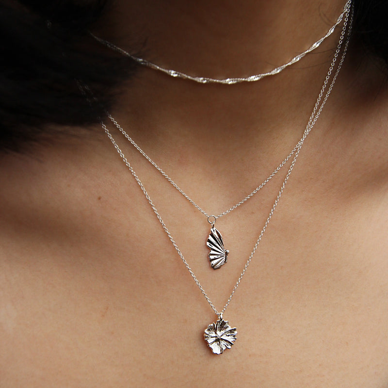 Close-up of a person wearing three layered silver necklaces. The shortest chain showcases a simple twisted design, the middle chain features a small fan-shaped pendant, and the longest chain displays bluboho's "larger wildflower necklace" in sterling silver, with a similar yet slightly larger fan-shaped pendant.