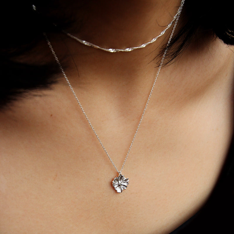 Close-up of a person wearing two delicate silver necklaces. One necklace is a simple chain, and the other is the "larger wildflower necklace" by bluboho, featuring a pendant shaped like a leaf with intricate detailing. The person is wearing a dark top, and their neck and collarbone are visible in the image.