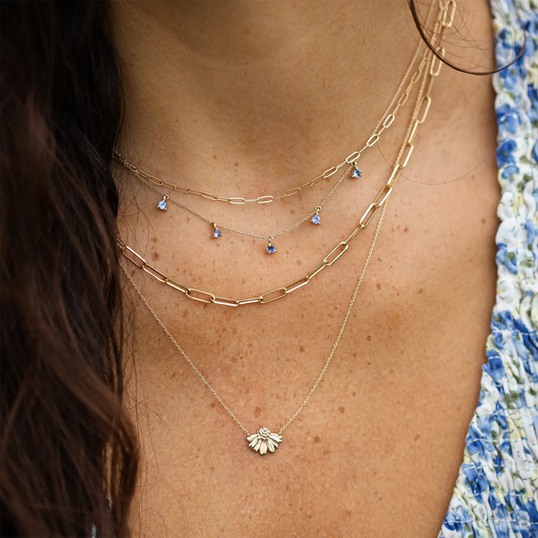 A person wears a wildflower tanzanite petal necklace - 14k yellow gold, tanzanite by bluboho. The shortest is a chain link necklace, the middle features small blue gems, and the longest has a daisy pendant. They are dressed in a blue and white floral pattern top.