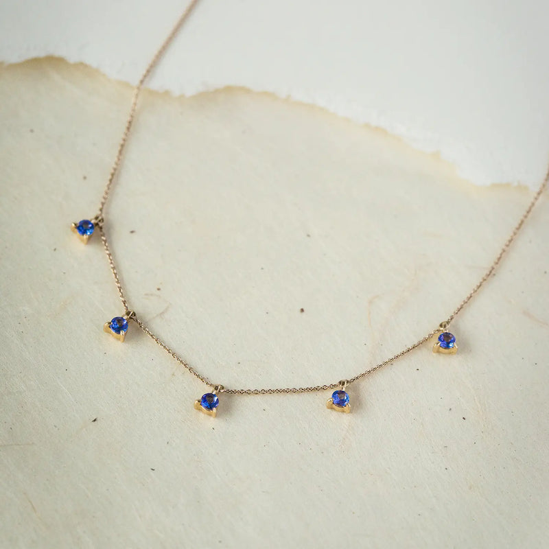 A delicate wildflower tanzanite petal necklace - 14k yellow gold, tanzanite by bluboho with evenly spaced blue gemstone pendants. The chain is thin and understated, with five small, round blue gems hanging from it. The necklace rests on a textured, neutral-colored surface.