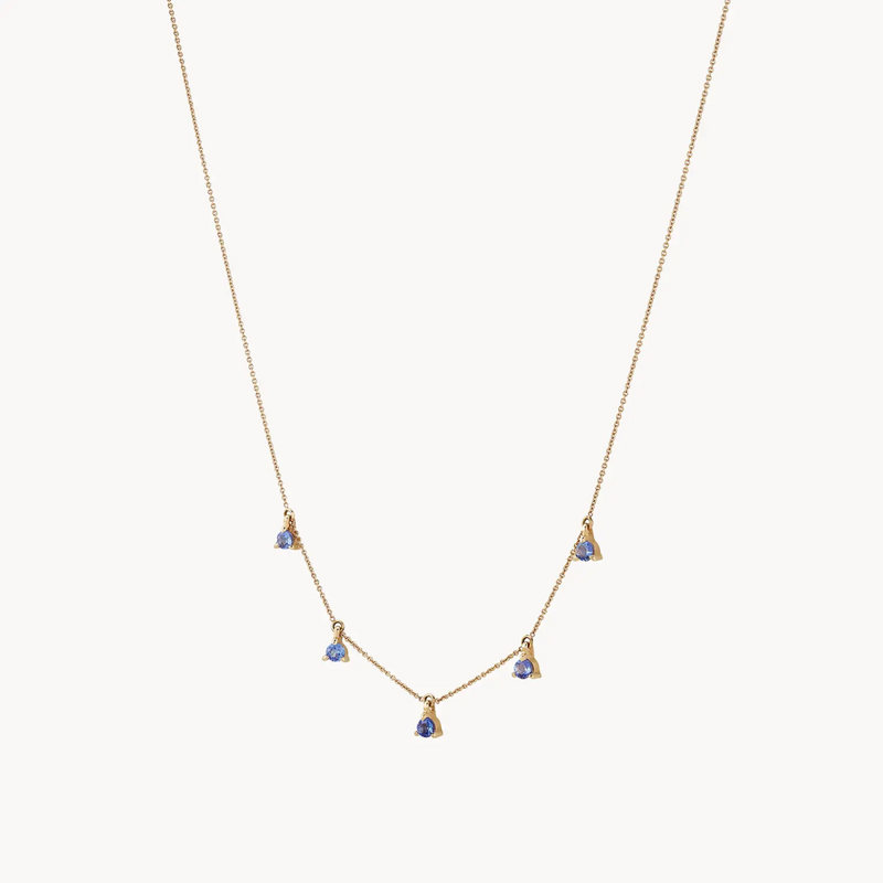 A delicate bluboho wildflower tanzanite petal necklace - 14k yellow gold, tanzanite with a fine chain, featuring five evenly spaced small blue gemstone pendants hanging along the front. The design is simple and elegant, suitable for a minimalist or sophisticated look. The background is plain white.