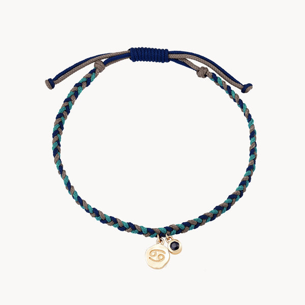 The bluboho cancer zodiac water element bracelet - 10k yellow gold, blue sapphire, cord features blue and green threads with an adjustable closure. The bracelet has two small charms: one is a gold charm with the Cancer zodiac sign, and the other is a small black bead.