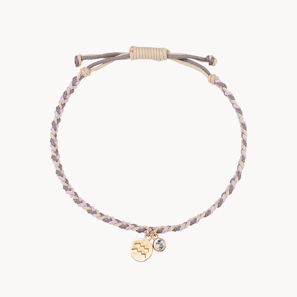 A braided bracelet made of light-colored cords with a sliding knot for adjustable sizing. Attached to the bracelet are two charms: one gold-colored charm with a zigzag pattern, and a small clear gemstone charm. The image is set against a plain white background. The product is the aquarius zodiac air element cord bracelet - 10k yellow gold, aquamarine, cord from bluboho.