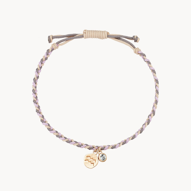 A braided bracelet made of light-colored cords with a sliding knot for adjustable sizing. Attached to the bracelet are two charms: one gold-colored charm with a zigzag pattern, and a small clear gemstone charm. The image is set against a plain white background. The product is the aquarius zodiac air element cord bracelet - 10k yellow gold, aquamarine, cord from bluboho.