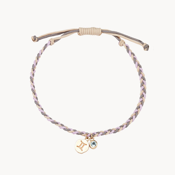 A braided bracelet made of light grey and lavender strands is pictured. It features two charms: a gold round charm with the Gemini zodiac sign and a small round blue gemstone. The bracelet has an adjustable sliding knot closure. This is the gemini zodiac air element cord bracelet - 10k yellow gold, aquamarine, cord by bluboho.