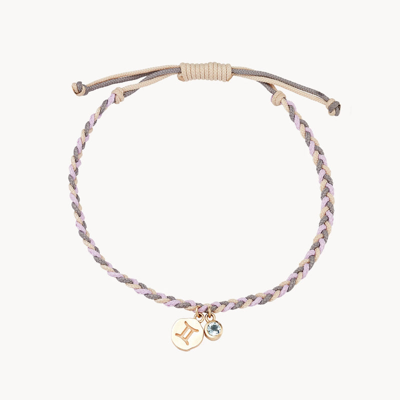 A braided bracelet made of light grey and lavender strands is pictured. It features two charms: a gold round charm with the Gemini zodiac sign and a small round blue gemstone. The bracelet has an adjustable sliding knot closure. This is the gemini zodiac air element cord bracelet - 10k yellow gold, aquamarine, cord by bluboho.