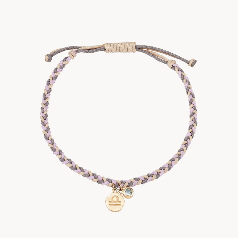 A bluboho libra zodiac air element cord bracelet - 10k yellow gold, aquamarine, cord featuring beige and lavender braided cords with an adjustable pull-tie clasp. The bracelet includes two small charms: one gold coin with the Libra zodiac symbol and another with a tiny round light blue gemstone.