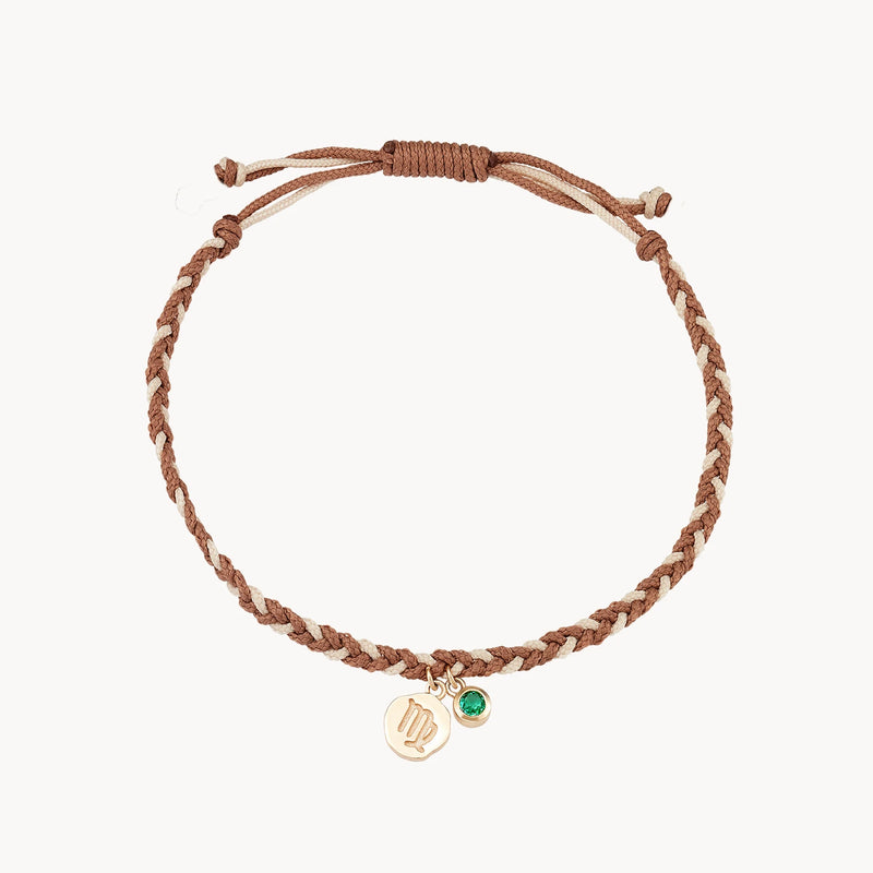 A brown braided string bracelet with two dangling charms: one gold circular charm with an embossed design and another green gem-like charm. The bracelet is adjustable with a sliding knot closure is the virgo zodiac earth element cord bracelet - 10k yellow gold, emerald, cord by bluboho.