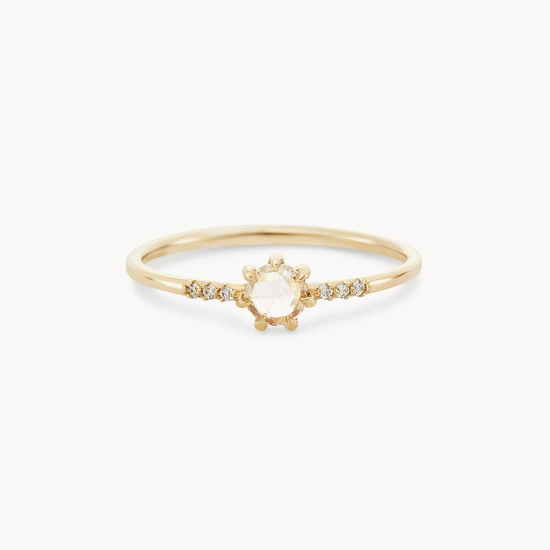 Product Sentence: The Supernova Ring by bluboho is a delicate 14k yellow gold ring featuring a central rose-cut white diamond set in a flower-shaped arrangement. The thin band is adorned with small round diamonds on both sides of the central gem, adding a touch of sparkle to its elegant and minimalistic design.