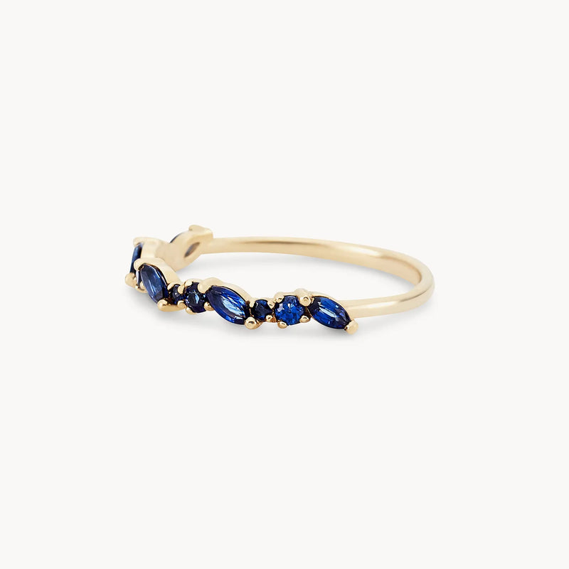 The "reverie band" by beloved by bluboho is a delicate 14k yellow gold ring featuring an elegantly thin band adorned with seven asymmetrically arranged, marquise-cut blue sapphires for a sleek and modern design.