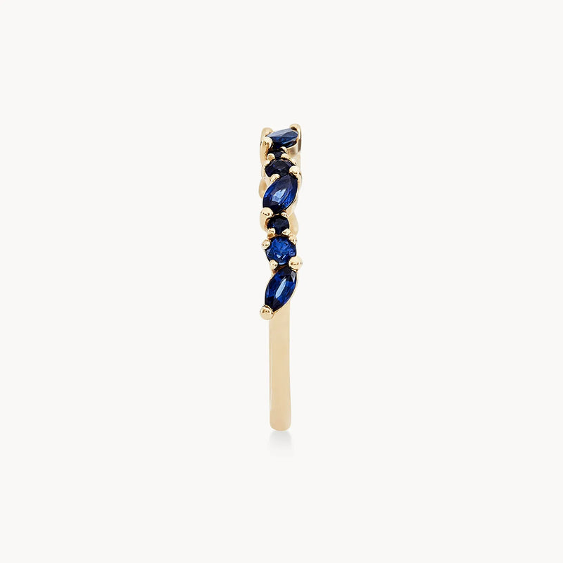 A single gold earring, known as the "reverie band" from beloved by bluboho, crafted in 14k yellow gold and featuring a vertical design adorned with clusters of deep blue sapphires. The minimalist and elegant appearance is enhanced by its slightly curved shape, against a plain white backdrop that highlights the exquisite craftsmanship.