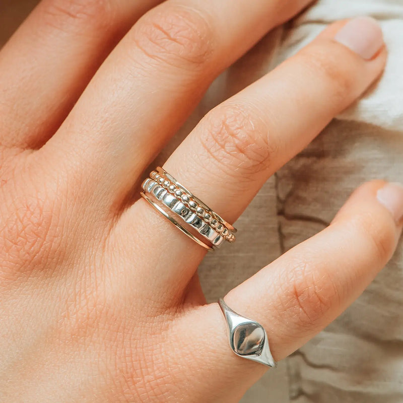 A close-up shot reveals a hand adorned with several rings. The middle finger showcases three stacked rings: one featuring a simple band, another with a beaded design, and a third embellished with an intricate pattern. On the ring finger rests the "peaks and valleys ring - sterling silver" by bluboho, distinguished by its unique shape. The nails are neatly manicured and natural.