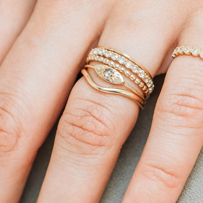 Close-up of a hand showcasing the "plain wave band - 14k yellow gold" from the brand Beloved by Bluboho. The rings adorning the ring and middle fingers include a band with a marquise diamond, a textured stackable ring, and several thin bands adorned with small diamonds. The skin appears smooth.