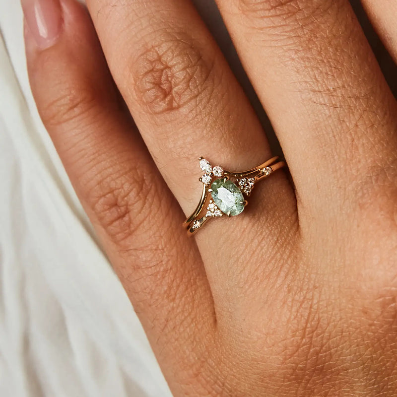 A close-up of a hand wearing the bluboho Mini Moonbow Ring in 14k yellow gold, featuring an elegant light green gemstone at the center surrounded by small diamonds. The ring boasts an intricate design, and the person's skin tone is light to medium. The background is a soft, textured white fabric.