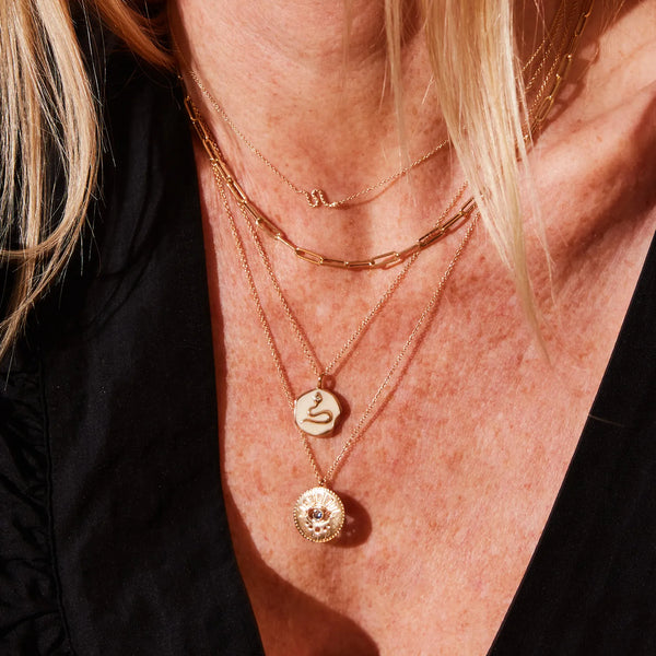 A person wearing layered gold necklaces with unique pendants from bluboho, including the revival snake enamel necklace in 14k yellow gold with a diamond and a coin-shaped pendant featuring a snake design. Another necklace has a medal-like pendant with intricate detailing, and the third is a simple, delicate chain. The person is dressed in a black top.