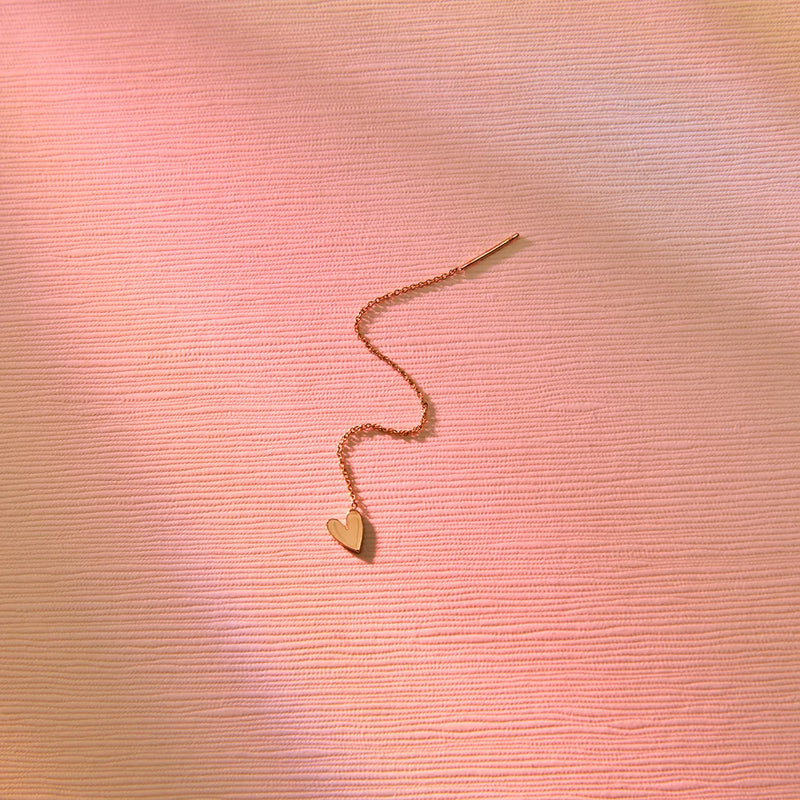 A delicate bluboho lovely heart threader earring - 14k yellow gold is placed on a textured pink surface. The soft lighting casts gentle shadows, highlighting the simplicity and elegance of the jewelry.