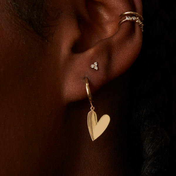 Close-up of an ear adorned with three pieces of jewelry: bluboho's "lovely heart sway hoop" crafted in 14k yellow gold, a small stud earring with three stones, and a gold ring in the helix. The person's hair is styled in braids, set against a dark and out-of-focus background.