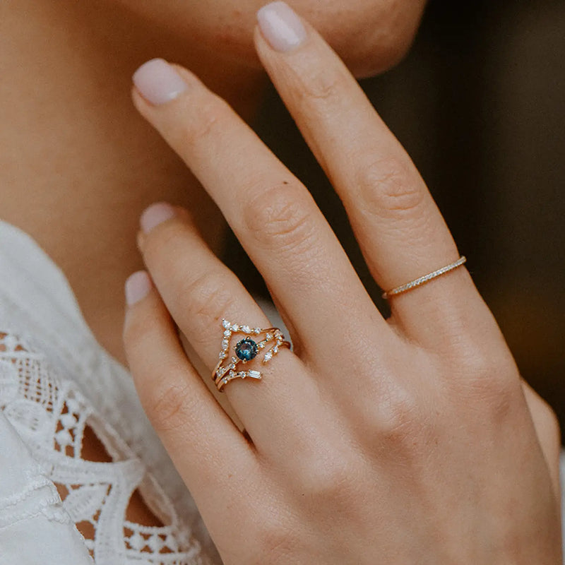 Close-up of a person's hand wearing two rings. One ring on the index finger is simple and thin, while the other on the ring finger is the Union of Bliss Ring by Beloved by Bluboho, featuring 14k yellow gold with white diamonds set around a dark central gemstone. The person is wearing a white lace top.