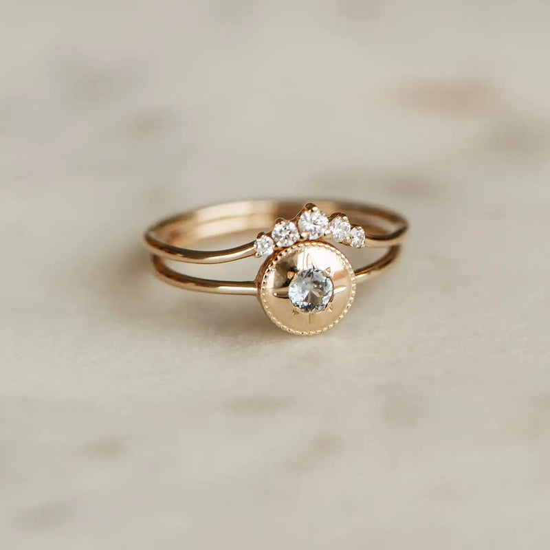 The Kismet Serendipity Ring by bluboho, crafted from 14k yellow gold and adorned with a central round white diamond surrounded by a crown of smaller diamonds, rests on a light-colored, textured surface. The ring boasts an elegant and delicate design with its double band.