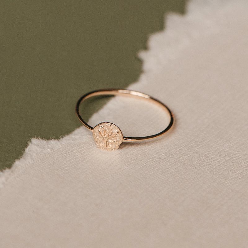 A delicate and elegant bluboho tiny tree of life ring in 14k yellow gold, gracefully displayed on a two-toned surface with one half being a textured white fabric and the other half a smooth green material.