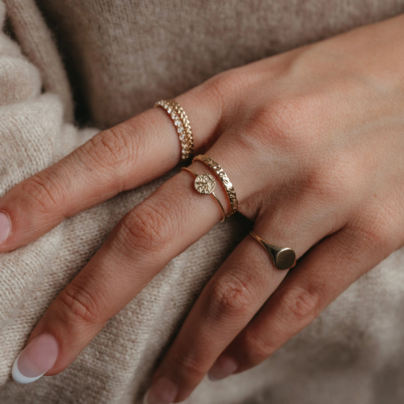 A close-up of a hand adorned with multiple gold rings on the fingers. The rings include designs such as a small medallion, a thin band with a tiny gemstone, and the tiny tree of life ring in 14k yellow gold by bluboho, featuring an intricate circular emblem. The person's nails are clean and natural, with a slight gloss.