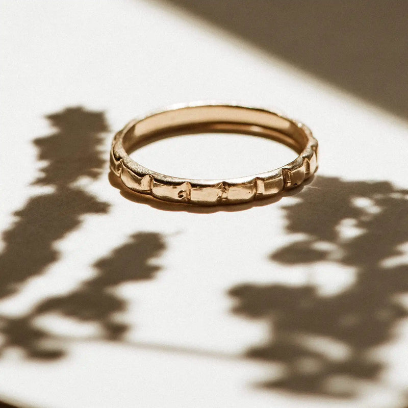 A bluboho peaks and valleys ring - 10k yellow gold lies on a surface with shadows of leaves and branches cast around it. The lighting creates a warm and natural ambiance.