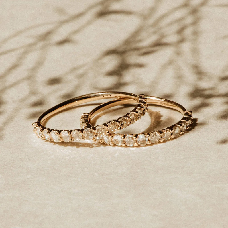 Two Moonglade rings, crafted from 14k rose gold and adorned with white diamonds by beloved by bluboho, are placed beside each other on a beige surface. Soft shadows and a blurred background with delicate branch-like silhouettes add an artistic touch to the composition.