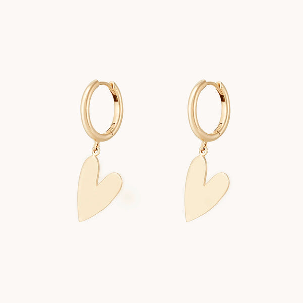 The lovely heart sway hoop - 14k yellow gold from bluboho is displayed. These smooth, round hoops each feature a polished, heart-shaped charm dangling gracefully below. The elegant simplicity of these earrings is accentuated by the plain, light-colored background.