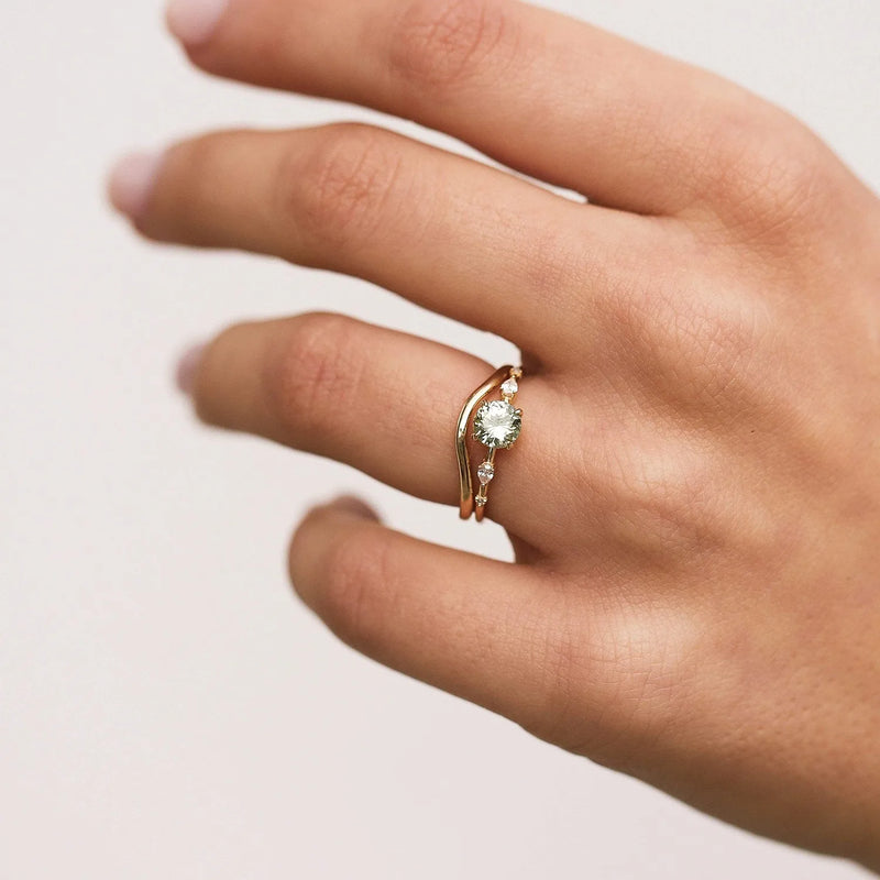 A close-up of a hand with a light skin tone showcases the "plain wave band - 14k yellow gold" by Beloved by Bluboho, featuring a large solitaire diamond. The ring is worn on the ring finger, and the nails are neatly manicured with a light pink polish. The background is plain and light-colored.