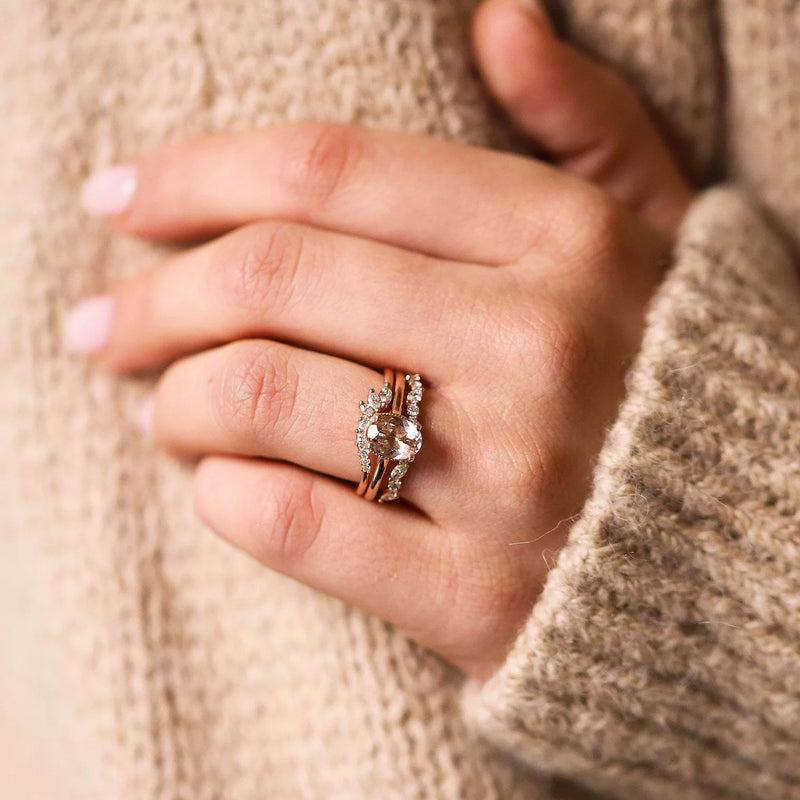 Close-up of a hand wearing the beloved by bluboho Incandescent band, a 14k yellow gold ring featuring a large central gemstone surrounded by smaller white diamonds. The hand rests against a textured beige clothing fabric, and the nails are painted light pink.