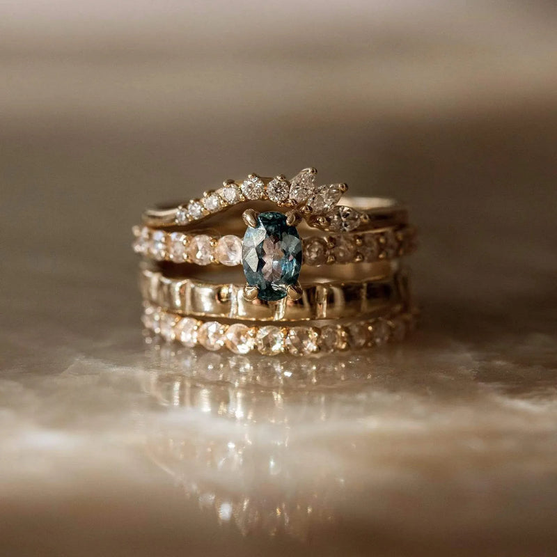 Here’s a close-up of a stack of elegant rings. The centerpiece ring is the Moonglade ring by beloved by bluboho, featuring a stunning blue-green gemstone set in 14k rose gold, flanked by small white diamonds, giving it the appearance of a tiara. Surrounding it are bands with various designs, including ones adorned with rows of diamonds and intricate details.