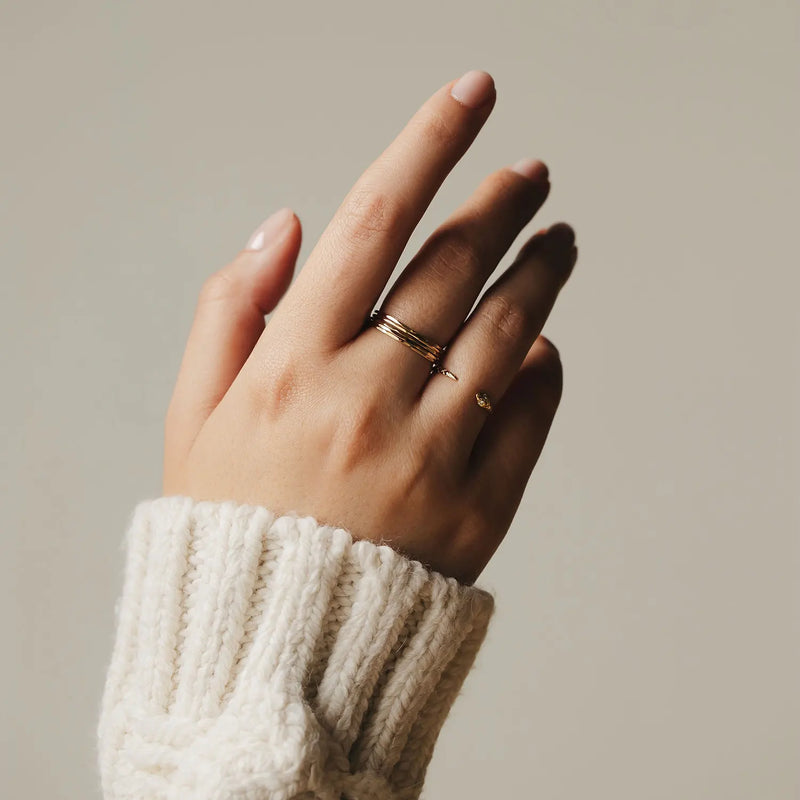 A hand, adorned with several Faceted Felicity Stacking Rings from bluboho in 10k yellow gold, wearing a chunky off-white knit sweater. The background is a plain neutral tone, accentuating the hand and its exquisite accessories.