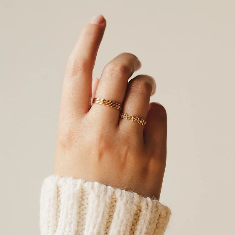 A hand with a cream-colored knitted sweater cuff is shown against a plain beige background. The hand wears two gold rings by bluboho: one is the plain jane stacking ring, a simple 10k yellow gold band on the middle finger, and the other is a delicate, decorative band with small interconnected circles on the ring finger.