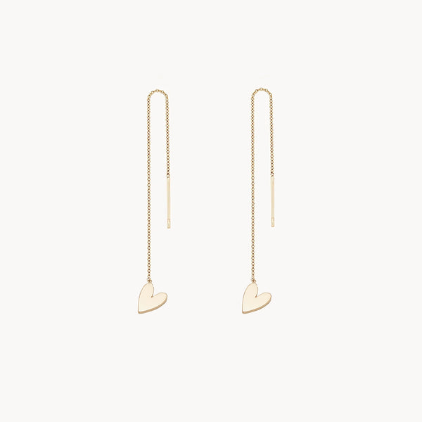 A pair of delicate gold threader earrings, each featuring a small heart-shaped charm at the end, displayed on a plain white background. The earrings have a thin chain that threads through the ear, creating a minimalist and elegant design. These are the lovely heart threader earring - 14k yellow gold by bluboho.