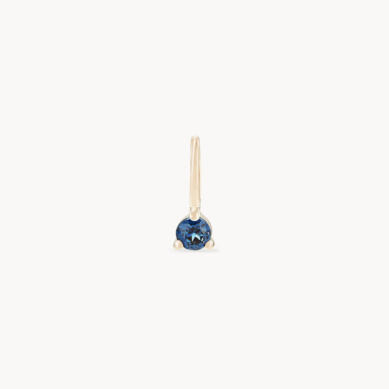 A close-up of the serendipity september birthstone charm by bluboho showcases a single earring featuring a small, deep blue sapphire set in a 10k yellow gold prong setting. The earring has a minimalistic design with a simple gold hook for threading through a pierced ear. The background is white.