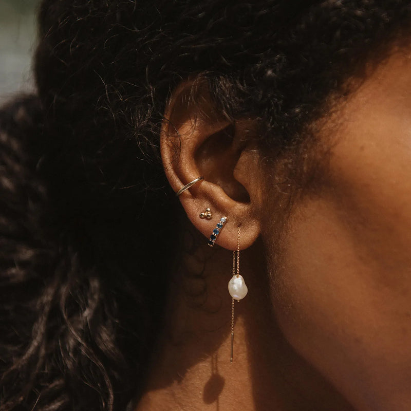 Close-up of a person's ear adorned with multiple earrings, including a small hoop, a stud with a sparkling gem, a blue and gold huggie, and the sofia perla threader earring from bluboho featured as the dangling pearl earring attached to a thin 14k yellow gold chain. The person's hair is styled in neat braids.