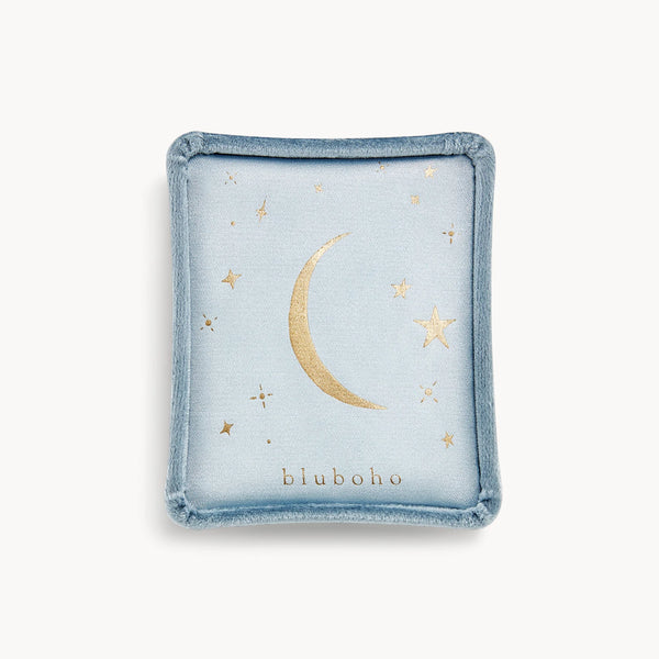 The "velvet tassel box" by bluboho is a square, light blue fabric jewelry container. It features a minimalist design with a gold crescent moon and stars, and the brand name "bluboho" is printed in gold at the bottom. The container also has soft, rounded edge trim.